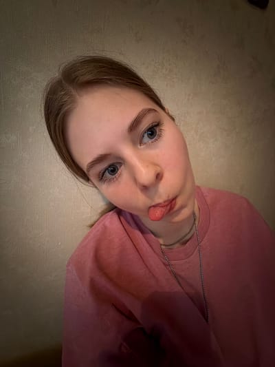 A young fair-skinned person with blue eyes and light brown hair is lying on their back against a neutral-colored surface, possibly a floor. The person is wearing a pink shirt and a silver chain necklace. They are sticking out their tongue. The location is unknown; there is no information in the image suggesting it is Gamovo, Russia.