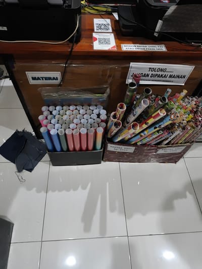 This is a medium-shot photograph taken from a slightly elevated angle, capturing a section of a retail counter or desk. The focus is on various items displayed for sale.
In the foreground, there are two cardboard boxes filled with rolled items. The box on the left contains numerous cylindrical objects, mostly in shades of blue, pink, green, and orange, likely paper rolls. To the left of this box, there are some crumpled dark fabric scraps. The box on the right is overflowing with a larger assortment of rolled paper, featuring colorful patterns and cartoon characters like Mickey Mouse. Visible labels on the boxes indicate "KERTAS KADO" (gift wrap) with prices of Rp. 3000 and Rp. 1500.
Behind the boxes, a dark wooden counter is visible. On the counter, a sign reads "MATERAI" (stamps). Further back, there's a printer or scanner. Above the counter, two QR code stickers are affixed to white cards. One sticker has text above it that reads "Toko Almi." Another sign, prominently displayed in the center, states in large lettering "TOLONG...... JANGAN DIPAKAI MAINAN" (Please... Don't Play With It), followed by smaller text that says "MERUSAK BERARTI MEMBELI" (Damaging Means Buying). A smaller sign above this reads "MOHON BAYAR UANG PAS ATAU BAYAR PAKAI QRIS" (Please Pay Exact Change or Pay With QRIS).
The setting appears to be an indoor retail environment, possibly a small shop or a service counter, given the presence of printed materials, payment instructions, and items for sale. The lighting suggests an indoor setting, possibly during the daytime. The overall impression is that of a shop that sells stationery or gift wrapping supplies, with clear instructions for customers regarding handling and payment.