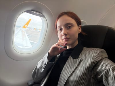 A female individual is seated in an airplane cabin, specifically identified as a Pegasus Airlines aircraft, positioned on an airfield at Sabiha Gökçen International Airport (SAW) adjacent to Tuzla, Turkey. The subject, with reddish-brown hair, is oriented towards the camera. Her right hand, adorned with pink nail polish, rests gently beneath her chin. She wears a black turtleneck garment overlaid with a black and white checkered blazer featuring solid black lapels. To her left, an oval aircraft window displays a white airplane wing in the immediate foreground, obscuring the engine. Beyond the wing, a yellow vertical stabilizer with the Pegasus Airlines logo is distinctly visible on the tarmac, alongside other aircraft and a cloudy sky. The ground visible through the window appears to have patches of snow or frost.