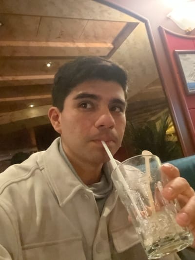 A man is drinking from a glass with a straw. He wears a light-colored jacket over a gray hoodie. The glass appears nearly empty, possibly with ice. He's looking at the camera with a serious expression. Behind him is a blurry interior scene with wood paneling and glimpses of a picture frame and greenery. The photo has a slightly rotated perspective.