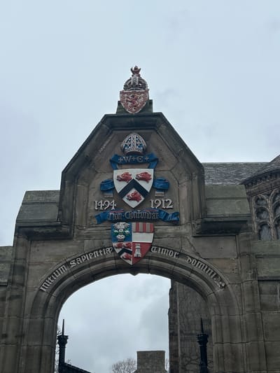 Image depicts a stone archway with elaborate heraldry above it. The archway features Latin inscriptions; "Amor Domini Initium Sapientiae". Above the arch is a heraldic display with multiple shields, including one with three boars' heads, and dates 1494 and 1912. The archway is part of a stone structure, likely a building. The sky in the background is overcast. Location: Likely within the University of Aberdeen, Scotland, United Kingdom.