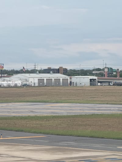 This image depicts a landscape in what appears to be San Antonio, Texas, United States of America. There is an airport with an American flag on top of the airport. The landscape includes airport runways and green space. There are buildings and industrial structures in the background, along with power lines and light poles. A highway and adjacent city street are running in the background. The sky is overcast.