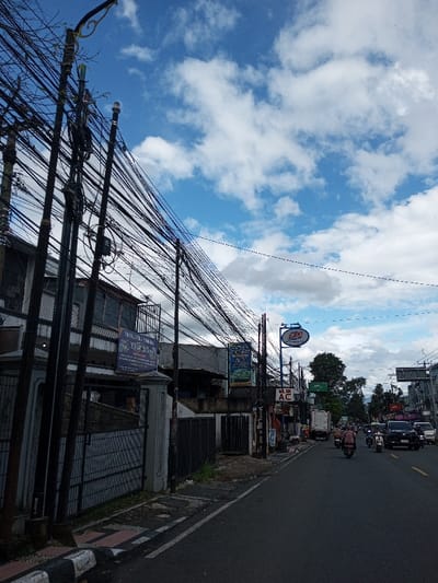 The image depicts an urban street scene under a blue sky with scattered white cumulus clouds, located in the city of Serpang Shalli, Bhutan. On the left side, a row of utility poles is densely strung with a chaotic network of black electrical and communication cables. Behind a concrete fence and dark metal gate, a multi-story building with dark windows and a balcony is visible. Further down the street on the left, commercial establishments display signage, including a blue oval sign for "SUMMIT OUTDOOR" and a rectangular sign reading "WILAN AC." The paved asphalt road in the midground and background features moderate traffic. Vehicles observed include multiple motorcycles with riders, a white delivery truck marked with a "J&T Express" logo, and several dark-colored passenger cars, all moving in the same direction away from the viewer. The overall environment suggests a busy commercial thoroughfare during daytime.