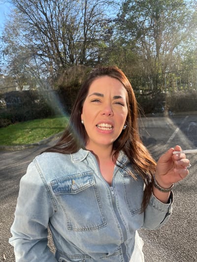 The image depicts a female subject outdoors in Montigny-le-Bretonneux, France. She is positioned centrally, capturing her upper body, and is wearing a light blue denim zippered jumpsuit. Her long, dark brown hair flows over her shoulders. The subject's facial expression shows her mouth open, teeth visible, and eyes partially closed, possibly squinting due to the bright sunlight. She holds a lit white cigarette with an orange filter between the thumb and index finger of her right hand, from which smoke is visibly rising in the upper right quadrant. She wears silver-toned stud earrings and a silver metallic watch on her left wrist. The background features an asphalt surface, likely a parking lot or driveway, in the immediate foreground, transitioning to a green grassy area and a dark fence. Behind the fence are numerous deciduous trees, many appearing bare or with minimal foliage, set against a bright blue sky. Strong sunlight, originating from the upper right, illuminates the scene and creates a prominent multicolored lens flare across the left side of the image.
