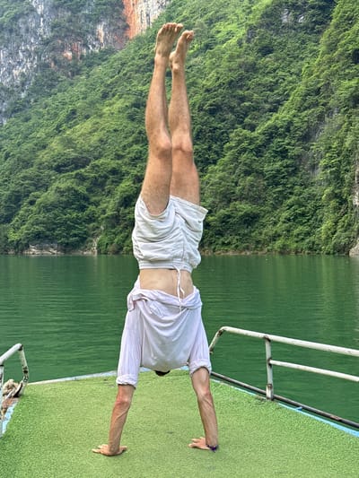 A person in white shorts and a white shirt is performing a handstand on a boat. The boat has green astroturf and a white metal railing. Behind the person, the backdrop is split; on the left is a steep cliff covered in dense green vegetation, and on the right is the still, green water of a river or lake. The location is Đồng Văn Commune, Vietnam, based on the landscape.