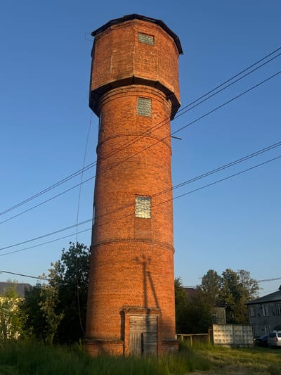 The image shows a tall, cylindrical brick water tower with a hexagonal top, situated in the village of Mostovik, Russia. The tower is constructed of reddish-orange brick and has several small, square windows with translucent glass panes visible along its height. A wooden door is present at the base of the tower. Overhead, multiple electrical wires traverse the frame. Surrounding the tower is a grassy area with trees and other buildings in the background, including what appears to be a residential building with grey siding and a fence. The sky is clear and blue, suggesting a daytime setting.
