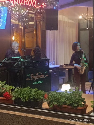 The image depicts a scene through a window, likely from a street, looking into a cafe or restaurant in Kislovodsk, Russia. A man with gray hair and glasses is seated at what appears to be an electronic keyboard, with a microphone in front of him. He is wearing a dark shirt and his chin rests on his hand, suggesting he is engaged in thought or listening. To his right, a woman with short dark hair, wearing a black top, is standing and playing a saxophone.
Prominently displayed on the equipment in front of the man is text that reads "КАФЕ Веранда" (Cafe Veranda) and "КИСЛОВОДСК" (Kislovodsk), confirming the location. The cafe appears to be dimly lit, with Christmas lights strung along the ceiling and a chandelier visible in the background. Behind the musicians, sheer curtains are drawn, and tables and chairs are partially visible. In the foreground, several potted plants with green foliage and red flowers are placed on the windowsill, suggesting an outdoor seating area or decorative elements. A digital timestamp in the bottom right corner reads "02.07.2026 20:25".