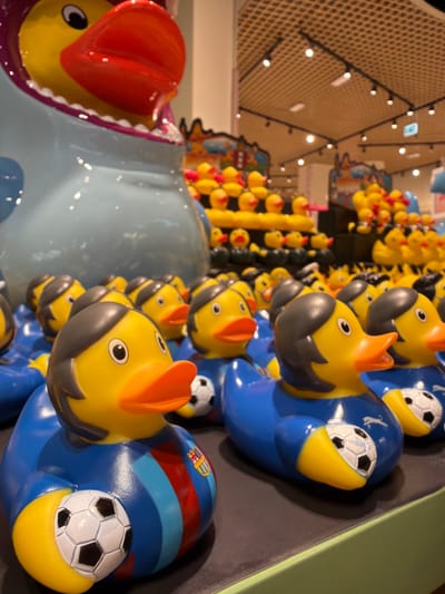 This media file shows a collection of yellow rubber ducks, some of which are dressed in blue and red striped outfits reminiscent of a soccer team's jersey. These ducks are adorned with miniature soccer balls on their beaks. In the foreground, a larger, light blue plush toy, possibly a shark or whale, is partially visible. It has a red cap or headpiece and a yellow duck head peeking out of its mouth. The background reveals a wall with a geometric pattern and numerous small lights, suggesting an indoor setting, perhaps a store or an arcade. The overall impression is playful and colorful, with a focus on toys. There is no indication of time of day, weather, or specific emotions. The location context provided is Fiumicino, Italy, but there are no explicit visual cues within the image to confirm this. No visible text is present in the image.
