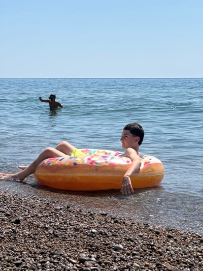 In Đurovići, Montenegro, on a sunny day with clear blue skies, a young boy is floating in the sea on a donut-shaped inflatable. He is lying on his back with his legs extended and resting on the inflatable, which is partially submerged in the shallow, clear water. His arms are relaxed, with one hand resting on the edge of the inflatable and the other slightly submerged. He is wearing yellow swim trunks. In the background, another boy, wearing a snorkel mask, is standing in the water and raising one hand. The foreground is dominated by a pebble beach, with small, wet stones visible where the water meets the shore. The sea extends to the horizon, meeting the sky.