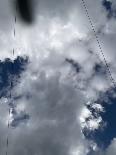 The image shows a view of a cloudy sky, likely taken from the ground looking upwards, with two thin, dark lines crossing the frame diagonally from the top left and top right towards the bottom center. These lines appear to be cables, possibly power lines or part of a transportation system. The sky is filled with a mix of dark and light grey clouds, with patches of deep blue sky visible between them. The clouds are varied in texture, with some appearing dense and others wispy. There is a large, dark, blurred mass in the upper left quadrant of the image, which could be a shadow or a closer object obscuring the view of the sky. The image does not contain any people, identifiable subjects, or specific objects that would indicate a particular activity or interaction. The scene is a natural skyscape. The location of Жостово, Russia, is provided as context, suggesting this is a sky view within or near that area.
