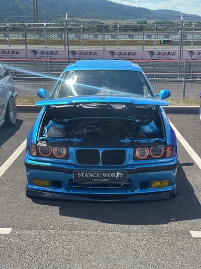 In Dospey, Bulgaria, a blue BMW E36 coupe with its hood open is parked on an asphalt surface marked with white lines, likely at a racetrack or car show. The car features orange halo headlights and a front license plate that reads "STANCE WORKS BULGARIA." In the background, a safety fence separates the parking area from a track with various sponsor banners, including "DIANA TYRES FOR YOU." Hills and a clear sky form the backdrop. The scene is brightly lit by sunlight, casting shadows on the ground.