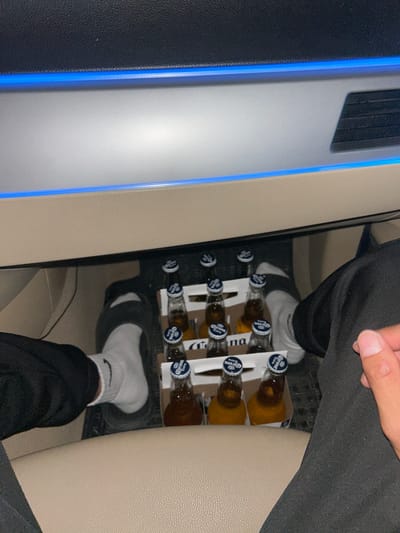 A six-pack of Corona Extra beer bottles is arranged in a white cardboard carrier. The bottles are mostly filled with amber liquid and have white and blue caps. Some of the bottles are slightly obscured by a person's hand and leg. The scene appears to be the interior of a vehicle, possibly a car or van, with light-colored leather seats. There is a black textured surface on the left, possibly part of the car's interior, with a blue illuminated accent. The image is taken from a high-angle perspective. The lighting suggests it might be daytime, but the exact time of day is not discernible. No specific weather conditions are indicated. The setting does not provide strong location cues beyond it being inside a vehicle. The visible text on the bottle caps reads "Corona Extra." The overall impression is casual, possibly indicating travel or leisure.