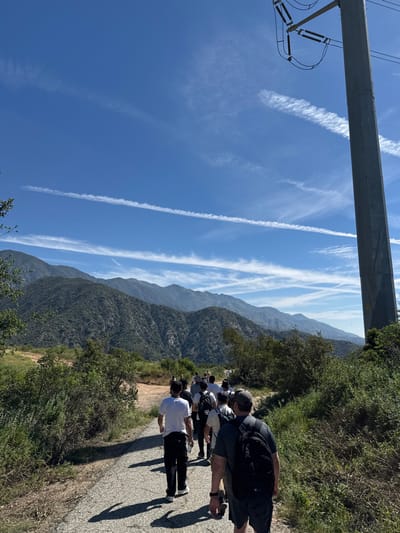 The image captures an outdoor scene in La Cañada Flintridge, United States of America, depicting a group of individuals walking along a paved path under a clear blue sky. A range of green, tree-covered mountains forms the backdrop, stretching across the horizon.
Approximately 10-15 individuals are observed from behind, walking away from the camera along a slightly winding, cracked grey path. They are dressed in casual attire including t-shirts, shorts, and long pants. Several individuals are carrying dark-colored backpacks. The individual closest to the camera wears a dark t-shirt, dark shorts, and a black backpack. Shadows from the walkers are cast on the path to their left, indicating bright sunlight from the right.
The path is bordered on both sides by dense green brush and scattered trees. On the far right of the frame, a tall, grey utility pole with exposed insulators and overhead power lines extends vertically, partially obscured by foreground foliage. The sky above is clear blue with multiple thin, white contrails or cirrus clouds visible, some appearing as distinct linear streaks.