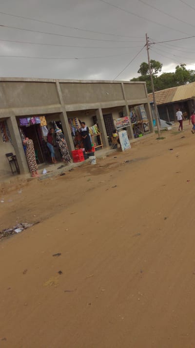 The image depicts a street in a low-income area, possibly in Masaka, Nigeria. There is a dusty dirt road occupying most of the frame's right side. The left side features a partially constructed, multi-story building with several open-fronted stalls. These stalls seem to be used for small-scale trading, with vendors selling items such as fabrics, groceries, and other goods. Some people are visible inside the stalls, apparently conducting business. Overhead power lines are present, and the sky is overcast with dull gray clouds. In the distance, additional buildings and trees can be seen. The general impression is that of a busy, informal commercial area in a developing urban environment.
