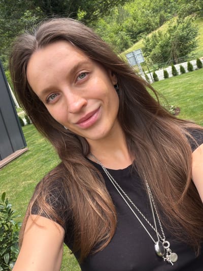 The media file is a portrait of a young woman with long, brown hair, taken outdoors in what appears to be a backyard setting. The woman is wearing a black top and a silver necklace with a pendant. Her face is turned towards the camera, and she has a slight smile. She has green eyes and light skin.
The background consists of a well-maintained green lawn, trees, and some fencing. The lighting suggests it is daytime, possibly late afternoon, with a pleasant outdoor atmosphere. The scene evokes a casual and relaxed mood. There is no visible text in the image. The location context provided is Bistrica, Montenegro, which aligns with the natural outdoor scenery depicted.