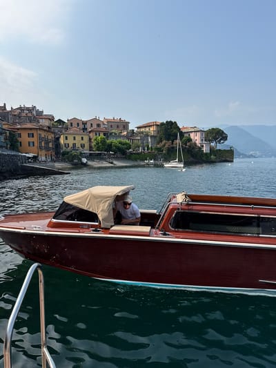 The image captures a scene in Varenna, Italy, focusing on a dark red wooden boat with a beige canopy. A man wearing a white t-shirt, sunglasses, and a cap is seated inside the boat, looking towards the right. The boat is docked in clear, dark green water, with a stainless steel railing visible in the foreground. In the background, a picturesque waterfront town with colorful buildings and a rocky shore is visible. Mountains rise in the distance under a clear blue sky with a few wispy clouds. A white sailboat is anchored offshore.