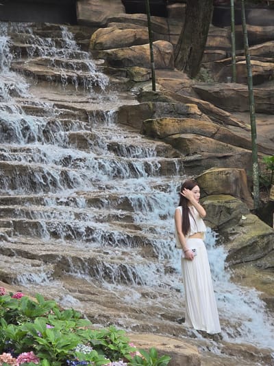 The image depicts a tiered waterfall with water cascading over rough, rocky surfaces. In the foreground, lush green foliage and clusters of pink and purple flowers are visible. A young woman, wearing a white sleeveless crop top and a flowing white maxi skirt, stands on a rocky outcrop to the right of the waterfall. She is holding a light pink smartphone in her right hand and appears to be adjusting her hair with her left hand. Behind her, bamboo stalks rise vertically against a backdrop of large, weathered rocks and a tree trunk. The scene suggests an outdoor natural setting, potentially a park or garden area within Xiaoshan District, China, known for its scenic landscapes and recreational facilities.