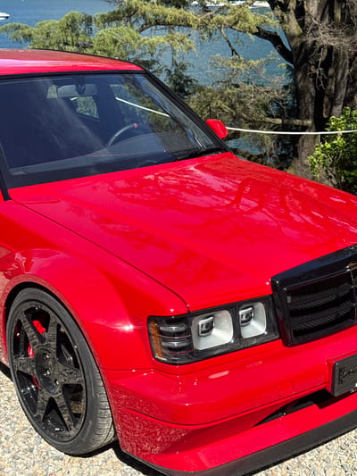 The image shows a close-up of a red vehicle, specifically the front right section including the hood, headlights, front fender, and front wheel. The car is parked on a gravel surface. A reflection of trees and water, potentially a lake, is visible on the hood of the car, hinting at a location near water and greenery. The black wheels have a multi-spoke design with red brake calipers visible behind them. The tire brand visible is Hankook. Based on the reflection and the gravel, it is likely that the image was taken near a waterfront or lakeside location, possibly in Como, Italy, given the mention of the city. No people are visible in the frame.
