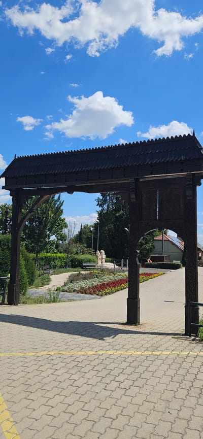 The image shows a large, ornately carved wooden gate in the foreground, framing a view of a park area with flowerbeds, benches, and a sculpture. The gate has a pitched, shingled roof and carved wooden posts. Beyond the gate, a pathway made of interlocking pavers leads through a garden with colorful flowers in reds, yellows, and whites. Two benches are visible in the park, along with a sculpture that appears to be made of light-colored stone. Trees and shrubs form a backdrop to the park. The sky above is blue with scattered white clouds. The overall scene suggests a pleasant outdoor setting in a public space. This location is identified as the gate to the Margit Vista in Cserszegtomaj, Hungary.