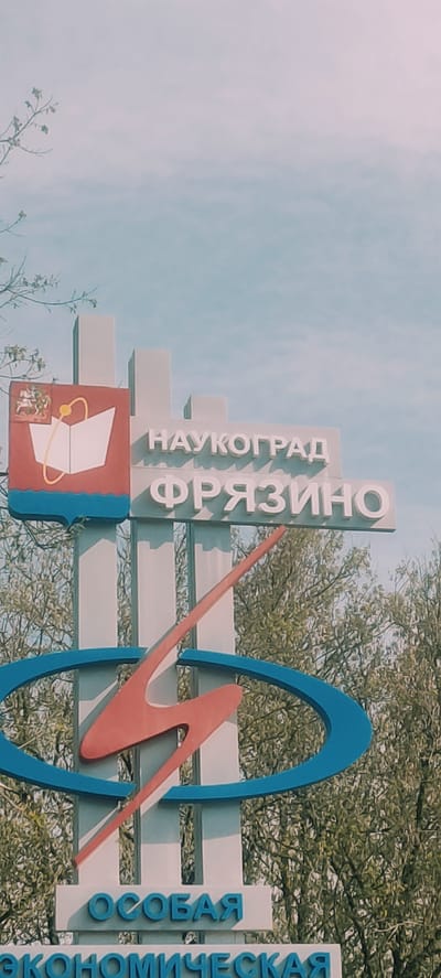 A sign in Fryazino, Russia, displays the city name in Cyrillic: "НАУКОГРАД ФРЯЗИНО". Below the text is a red square with a blue border containing a white book graphic surrounded by a gold atom model and a small emblem at the bottom. Beside this is a blue oval intersecting a red stylized 'M'. Beside this is the Cyrillic text "ОСОБАЯ ЭКОНОМИЧЕСКАЯ". The sign is located outside amidst foliage and a partially cloudy sky.
