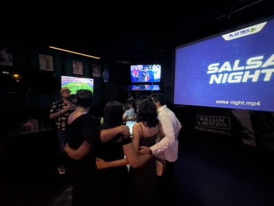 This image captures a lively evening scene inside a dimly lit entertainment venue in Colombo, Sri Lanka. A group of adults is gathered, engaging in social interaction, possibly dancing or socializing closely.
In the foreground, several individuals are seen from behind or partially, with their arms around each other, suggesting dancing or close conversation. One man in a fedora hat and a patterned short-sleeve shirt stands to the left, looking towards the group.
The venue is equipped with multiple large screens. A prominent screen on the far right displays "SALSA NIGHT" in large white letters, with a "PLAYTRIX" logo in the top right corner and "salsa night.mp4" at the bottom, indicating a dance event or themed night. Below this screen, a white sign for "WILLIAM LAWSON'S" whisky is visible, stating "NO RULES. GREAT SCOTCH."
To the left of the main screen, another large screen is showing a football (soccer) match on a green field. Above this, a smaller screen displays an indoor scene with a few individuals, one wearing a jersey that appears to say "ITALIA," likely showing a sports-related interview or pre-game coverage.
The walls are dark, adorned with framed pictures or posters. A bright, thin yellow light strip runs across the upper part of the wall, adding to the ambient lighting. The overall atmosphere suggests a vibrant and engaging social setting, likely a bar or club, taking place at night. The focus on screens showing both sports and a "Salsa Night" indicates a diverse entertainment offering.