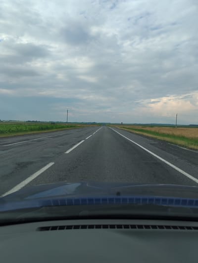 The image depicts a view from the interior of a vehicle looking out at a rural road in the Урыцкі сельскі Савет, Belarus. The road is paved and appears to have some sections with visible wear and tear. White lines mark the lanes of traffic, and the road stretches into the distance, seemingly toward a horizon where fields of crops and vegetation are visible. The sky is overcast with a mix of grey and white clouds. Utility poles are present along the side of the road. The foreground shows the hood and dashboard of the vehicle from which the photo was taken. There are no people or animals visible in the image.