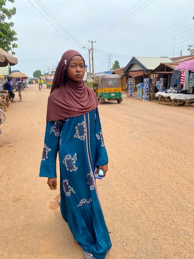 A young woman wearing a dark teal abaya with ornate gold and white patterns stands on a dirt road in Barakin, Nigeria. She is also wearing a maroon hijab. She holds a smartphone in her right hand and her left hand is outstretched. The road appears to be a marketplace with various stalls lining the left side of the frame. These stalls display goods such as clothing, electronics, and other wares. A colorful three-wheeled auto rickshaw is parked on the side of the road. In the background, there are power lines and a hazy sky, suggesting it might be daytime with overcast weather. People can be seen milling about in the distance. The scene depicts a bustling marketplace environment.