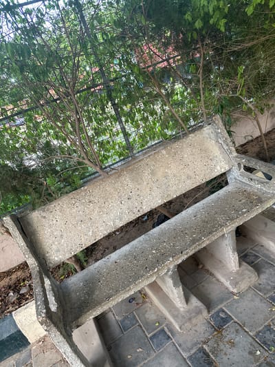 An outdoor scene in Batagarawa, Nigeria, features a weathered concrete bench situated on a paved walkway. The bench, composed of rough-textured concrete with visible aggregate, shows signs of age and use. It is positioned in front of a dense green foliage, likely a hedge or shrubbery, which forms a backdrop. A metal fence or trellis is partially visible behind the foliage. The ground around the bench is paved with rectangular tiles in varying shades of grey and black, interspersed with some dried leaves and debris. The lighting suggests it is daytime, possibly overcast. No people are present in the image.
