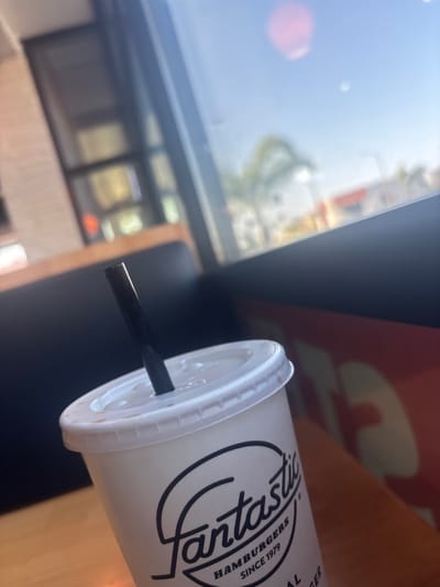A slanted view shows a fast-food drink cup with a black straw inserted through the lid. The cup has the "artastic" logo and states "hamburgers since 1979". The background is blurred, featuring a window with a light blue sky and glimpses of nearby buildings, plus some interior elements. The overall image composition and lighting suggest a casual, possibly outdoor or in-vehicle, setting.