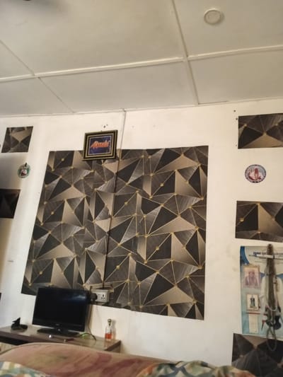 The image depicts an indoor scene, likely a room in a residential building in Ipota, Nigeria. The walls are painted white and feature a geometric patterned wallpaper in shades of grey, black, and gold. Several framed pictures and circular stickers adorn the walls. A framed picture with the word "Asante" is prominently displayed. A small television set is placed on a wooden desk below the wallpaper, next to a power outlet. Other objects on the desk include a bottle of liquid, what appears to be sunglasses, and a small black object. Part of a bed with a patterned comforter is visible in the foreground. The ceiling is white and appears to be made of suspended panels. The overall impression is that of a simple, decorated living space.