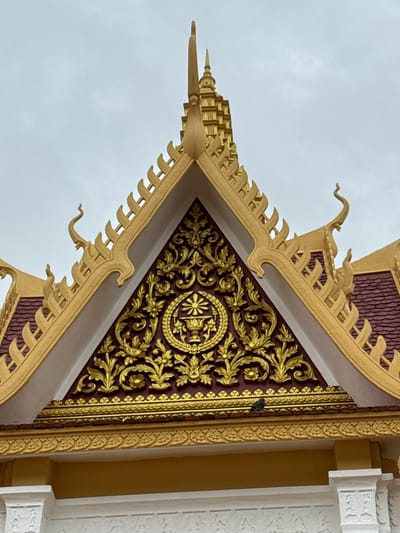 A close-up, low-angle shot captures the ornate golden roof of a Cambodian structure, likely a temple or royal building, against a pale, overcast sky. The roof features intricate gilded patterns and decorative finials, with a prominent triangular pediment adorned with elaborate floral motifs and a central circular emblem. Below the pediment, a tiled section of the roof is visible, colored a deep maroon. A single pigeon is perched on the edge of the roof structure, adding a small, living element to the otherwise static scene. The architecture suggests a location within Phnom Penh, Cambodia, a city known for its many Buddhist temples and historical buildings, particularly in the Khan Daun Penh district which houses many of the country's most significant landmarks.