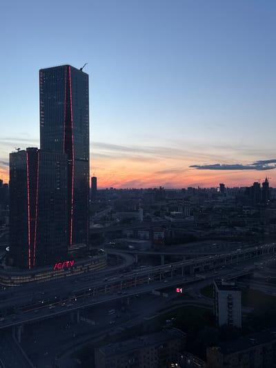 The image is an aerial view of a cityscape at dusk, dominated by two modern skyscrapers with prominent red lighting on their facades. The skyscraper on the left, taller and thinner, features vertical red lighting strips. To its right and slightly shorter, is another skyscraper of similar design. Below and surrounding the skyscrapers is a complex network of elevated highways and roads, with visible traffic including cars and buses. The city stretches into the background, characterized by a dense collection of buildings of varying heights. The sky exhibits a gradient of colors, transitioning from a clear blue at the top to a warm orange and pink hue near the horizon, indicating sunset. A few wispy clouds are scattered across the sky. In the far distance, other tall buildings and construction cranes are visible. The overall scene depicts a vibrant urban environment at the end of the day. The location of Туристический Пансионат "Клязьминское водохранилище", Russia, is not identifiable within this image; the scene depicts a large, modern city center.