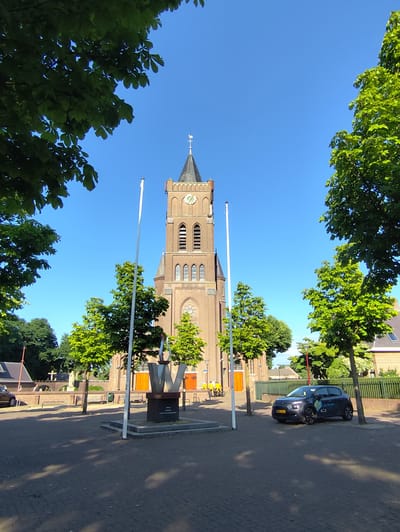 This is a photograph taken in Schaijk, Netherlands, depicting a church tower and its surroundings on a clear, sunny day. The time of day appears to be late morning or early afternoon, suggested by the bright sunlight and distinct shadows cast on the paved ground.

The main subject is the brick church tower, which has a clock face visible near its peak. A weather vane tops the spire. In front of the church, a modern sculpture resembling a stylized letter "W" with flames is situated on a dark pedestal. The pedestal has some text on it, but it is not clearly legible in the image.

The scene is framed by lush green trees with dense foliage, characteristic of a warm season, likely spring or summer. Two tall white flagpoles stand in the foreground, one to the left of the sculpture and one to the right, though no flags are visible.

A dark gray car is parked on the right side of the image. It has some decals on its side, one of which reads "Get the job!". Behind the car, a green fence runs along the property line. In the background, other buildings and trees are visible, contributing to the impression of a small town or village setting.

To the left of the church, a cemetery or memorial area is suggested by a cross and some statues. A few people are visible in the background near the cemetery, and two individuals on bicycles in bright yellow jackets are near the church entrance. The overall mood of the image is peaceful and serene, highlighting the architectural beauty of the church and the vibrancy of the surrounding nature.