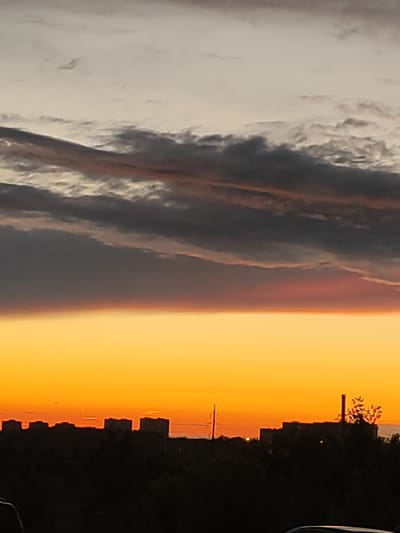 This is a vertical, medium-shot photograph of a sunset over a city. The sky dominates the frame, with a gradient of colors from pale grey at the top to a vibrant orange at the bottom. Dark, wispy clouds are layered across the upper portion of the sky, creating a dramatic contrast with the brighter hues of the sunset. The lower third of the image features a silhouetted cityscape with trees in the foreground. Several buildings, appearing to be apartment blocks, are visible along the horizon. A prominent industrial smokestack stands to the right of the frame, partially obscured by the silhouette of trees. A single, thin pole with wires extends upwards from the cityscape on the left. The bright orange glow of the setting sun is visible just above the silhouetted buildings, casting a warm light. The time of day is clearly sunset, and the weather appears clear with scattered clouds. The overall mood is serene and a bit dramatic due to the intense colors and cloud formations. The location is identified as Shchyolkovo, Russia. There is no visible text in the image.
