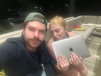 A couple is pictured upside down, likely a selfie. The woman, with blonde hair and several rings, holds a silver laptop with an Apple logo. The man, wearing a dark jacket and a cap, has a beard. They appear to be relaxing outdoors, possibly on a cushioned seating area. The background includes what appears to be outdoor furniture and dark surroundings. The lighting is somewhat dim.