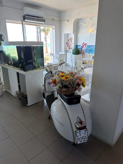 A white Vespa scooter, decorated with a basket of colorful flowers, is parked indoors in what appears to be a cafe or restaurant in Vallecrosia al Mare, Italy. The scooter is positioned in the foreground, facing away from the viewer, with its license plate clearly visible, reading "CT 7 7055". Behind the scooter, a large aquarium filled with fish and aquatic plants sits on a white stand. Further in the background, tables and chairs are set up, suggesting a dining area. A mural depicting figures in beach attire adorns the wall. The room is illuminated by natural light streaming in from a large window that offers a view of palm trees and the sea. An air conditioning unit is mounted on the ceiling. The floor is tiled.