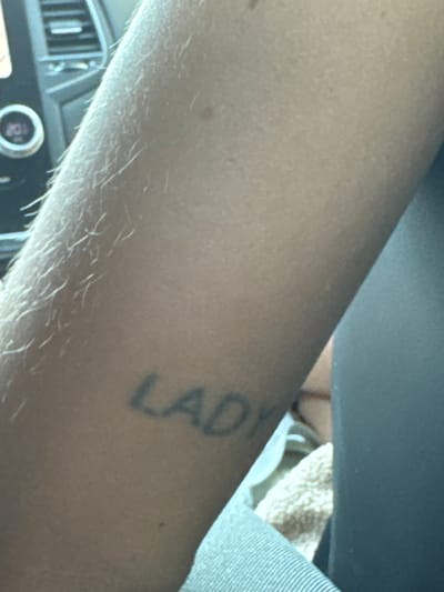 The image shows a close-up of a person's forearm with a tattoo of the word "LADY" in black ink. The tattoo is on the inner side of the forearm, appearing slightly faded. The skin is fair and has visible fine hairs. In the background, out of focus, a car's interior is partially visible, including a dashboard with vents and a circular dial. The lighting suggests it is daytime, possibly indoors or in a shaded area. There are no recognizable landmarks or indications of a specific location within Donja Cijevna, Montenegro. The image is framed to focus on the tattoo.