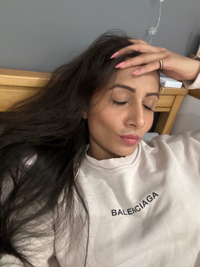 A female individual with long, dark hair, light-to-medium complexion, and closed eyes is positioned in a medium close-up indoors. She wears a light-colored crew-neck sweatshirt with the word "BALENCIAGA" printed in dark letters across the chest. Her right hand, adorned with pink nail polish and a gold-colored ring on the ring finger, rests on her forehead. Subtle makeup is visible on her eyelids, eyebrows, and lips, which are coated with a pink gloss. A thin, silver-colored hoop earring is visible in her right ear. Behind her, a wooden headboard is visible, above which is a plain grey wall. To the right of her head, a section of a wooden surface, possibly a nightstand, and a string of clear decorative lights are partially visible. The location within the city of Winslow, United Kingdom, cannot be determined from the image.