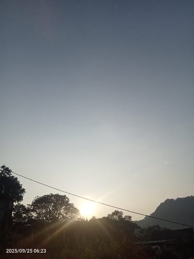The image depicts a sunrise scene in Quebrada del Medio, Venezuela, recorded at 06:23 on September 25, 2025. The sky transitions from a deep blue-grey at the top to a brilliant light yellow-white at the horizon. The sun, a bright white orb with prominent lens flare, is situated in the lower-middle portion of the frame, partially obscured by the dark silhouettes of dense trees. A dark utility wire spans horizontally across the lower third of the image, passing directly in front of the sun. In the background to the right, a dark, undulating mountain range is visible, heavily silhouetted against the brightening sky. No individuals or active subjects are present. The foreground consists of dark, indistinct foliage and what appears to be part of a structure or roofline below the trees on the left.