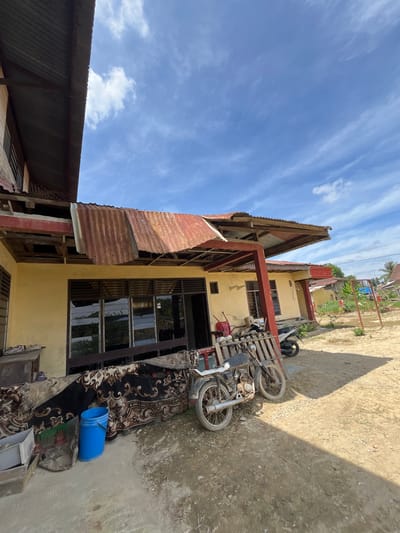 This media file depicts an outdoor scene, likely in a residential area of Kota Kuala Simpang, Indonesia. The image is oriented vertically, with the sky occupying the top portion and the ground and buildings in the lower part.

The main subjects are a partially constructed or dilapidated building and two motorcycles. The building features a yellow-painted wall with dark-framed windows. A corrugated metal roof is partially attached to one side of the building, with some sections appearing damaged or missing. Red metal beams are visible supporting parts of the structure. Adjacent to the building, two motorcycles are parked on a dirt ground. One motorcycle, closer to the building, is a light color with spoked wheels and appears to be on a wooden pallet or stand. Another motorcycle is partially visible further away.

On the ground near the motorcycles and building, a patterned blanket or rug is laid out. A bright blue plastic bucket is placed on top of the blanket. The ground itself is a mixture of dirt, gravel, and some sparse vegetation. In the background, more buildings and palm trees are visible under a bright blue sky with scattered white clouds. The sunlight casts a distinct shadow on the ground from the building, suggesting it is daytime, likely mid-morning or afternoon.

There are no people visible in the media file. The scene appears somewhat cluttered with the objects and the state of the building suggesting a potentially informal or developing area. The lighting and sky indicate fair weather. No visible text is present in the image.