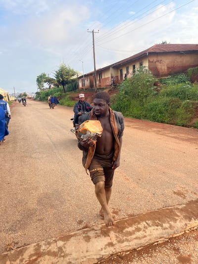 In Gembu, Nigeria, a bare-chested man walks on a paved road carrying a tan plastic jerrycan and a piece of cloth. He is wearing brown shorts with visible lettering and appears to be barefoot. Behind him, several other people are on motorcycles, including a man in a black jacket and white cap, and another person wearing a blue garment. The background consists of a row of buildings with corrugated metal roofs and lush green vegetation. The sky is cloudy.