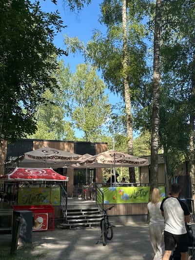 The image captures a sunny day scene in Dubrovitsy, Russia, likely near the Church of the Holy Virgin. In the foreground, a red and white ice cream stand with the brand "Unilever" is visible, featuring a prominent red heart logo and Russian text, "УРА!!! МОРОЖЕНОЕ!" (Hooray!!! Ice cream!). Beside it, a green banner with yellow lettering reads "ЛЕТО" (Summer). A black bicycle is parked near the stand. In the mid-ground, a casual outdoor seating area with umbrellas is set up outside a building with a wooden facade. Several people are seated at tables, enjoying their time. A couple, a man and a woman, are walking away from the camera towards the right, the man wearing a white and black shirt and black shorts, and the woman in a white top and light-colored pants. Behind them, part of a stroller is visible. The background is dominated by tall trees with green foliage, suggesting a park or natural setting within the urban environment. The sky is clear and blue.