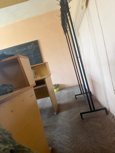 The image shows an interior view, potentially a classroom or office space, in Dolna Mitropolia, Bulgaria. The walls are light orange, and the floor is covered in a herringbone pattern of light brown tiles. Visible in the room are several wooden cabinets or shelving units, a black chalkboard with handwriting in white chalk, and a black metal clothing rack attached to the wall near the ceiling. There are no people explicitly visible, but there may be an individual partially visible in the lower right corner of the image.