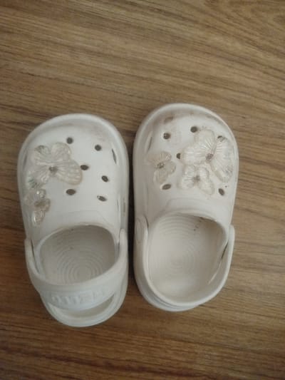 A pair of white children's Crocs are pictured lying on a wooden floor. The Crocs have decorative, translucent butterfly charms attached to the top, with one larger butterfly on the left shoe and two smaller ones on the right. The shoes appear to be slightly worn and dusty, suggesting they have been used. The wooden floor has a natural grain pattern. The overall scene is domestic and casual. No activity or event is taking place; it is a static image of the shoes. The lighting is neutral, and no specific time of day or weather conditions are apparent. There is no visible text in the image. The location is Criuleni, Moldova.
