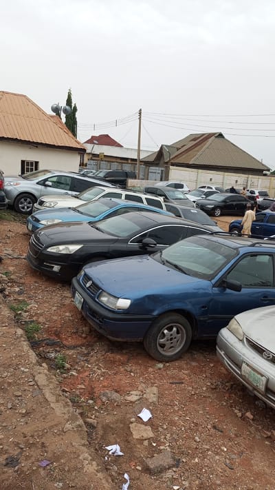 The image depicts a used car lot in Kawo, Nigeria, on an overcast day. Several rows of various cars are parked on a dirt lot with gravel and scattered debris. The vehicles appear to be predominantly sedans, including Peugeot, Toyota, and other models, showing signs of age and use. Buildings with corrugated metal roofs and painted walls form the backdrop, suggesting a residential or commercial area. A single individual is visible wearing a traditional outfit, walking near the parked vehicles. Overhead power lines are also visible. The overall scene suggests a used car dealership or storage area in a working-class neighborhood.