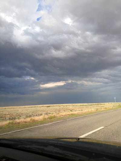 The image depicts a wide, paved road cutting through a flat, grassy plain under a dramatic, cloudy sky. The foreground is dominated by the dark dashboard of a vehicle, suggesting the photo was taken from inside a car. The road itself is asphalt, with a white line marking the edge. Beyond the road, a vast expanse of dry, golden-brown grass stretches to the horizon, dotted with occasional low shrubs. The sky is filled with heavy, dark grey storm clouds, with patches of lighter clouds and a hint of blue sky visible in the upper left. In the distance, a thin, pale band of light suggests a break in the clouds or the sun trying to penetrate the gloom. A solitary white signpost with a blue symbol is visible on the right side of the road, near the horizon. The overall impression is one of an approaching storm in a vast, open landscape. The location is specified as Khulkhuta, Russia.
