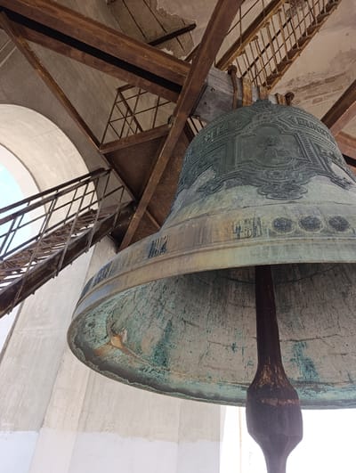This media file depicts a close-up, low-angle view of a large, weathered bronze bell, likely in a bell tower or belfry. The bell is green with verdigris patina and exhibits some rust and discoloration, indicating age and exposure to the elements. Intricate relief carvings adorn the upper portion of the bell, including what appears to be a religious figure in a central medallion, surrounded by decorative borders and inscriptions. Below this, there is a band with Cyrillic inscriptions and some circular markings.

The bell is suspended from a robust metal structure composed of large, rusty I-beams and supporting wooden elements. To the left, a portion of a white, arched structure is visible, suggesting the architectural context of a church or historical building. A metal staircase with a grated surface ascends diagonally to the right behind the bell, also showing signs of rust and wear, leading further up into the unseen structure. The overall impression is one of historical significance and enduring, perhaps slightly decaying, grandeur.

The lighting suggests it is daytime, likely sunny, given the brightness and contrast in the image. The scene evokes a sense of stillness and perhaps solemnity, with the bell presented as a silent sentinel. While no people are visible, the presence of the bell and the infrastructure implies a functional space, possibly visited by people for religious services or historical interest. The location context provided is Kitskany, Moldova. The visible text on the bell is in Cyrillic, but it is too distorted and partially obscured to be definitively read.
