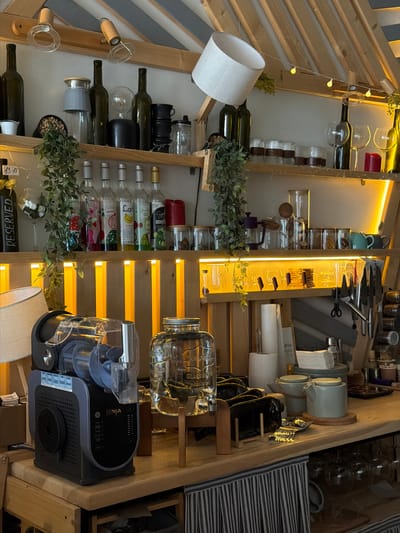 This image depicts the interior of a cafe or bar, likely in Volochysk, Ukraine, characterized by its rustic and wooden aesthetic. The scene is densely populated with various items displayed on shelves and a countertop.
On the upper shelves, an assortment of glass bottles, some dark green and others clear, are arranged. Several are full, while others appear empty. Among the bottles, there are jars containing what looks like dried herbs or spices, and a few decorative items like a lampshade, a light bulb, and a red candle. Hanging greenery drapes from the shelves, adding a natural element.
The middle shelf features a row of clear glass bottles, some with colorful labels indicating flavored syrups, such as "Strawberry," "Lychee," "Banana," and "Mojito." Next to these are wine glasses and more jars with dried contents.
The lower shelf and countertop area are even more cluttered. A black and gray Ninja food processor is prominently positioned on the left. To its right, a large glass dispenser with a spigot sits on a wooden stand. Various kitchenware, including teapots, mugs, and stacks of napkins, are visible on the countertop. Below the countertop, a striped fabric hangs, partially obscuring shelves with glassware.
The environment is lit by warm, artificial lighting, creating an inviting atmosphere. The overall impression is that of a well-stocked establishment with a focus on drinks and possibly light refreshments, with a strong emphasis on decorative elements that contribute to a cozy ambiance. There are no people visible in the image.