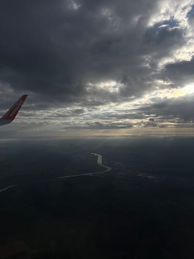 The image, taken from an airplane, shows a view of clouds, ground, and a wing. The lower-left corner includes part of an airplane wing with the word "easyJet" visible. The left side of the image is dominated by dark clouds, with glimpses of lighter areas and some blue sky. A strip of light, possibly sunlight filtering through the clouds, stretches across the middle of the frame. To the right of this light strip is a dark area which is the ground, with a river winding through the landscape. It is not possible to pinpoint the exact location to the city of Berg, Luxembourg using the current information.