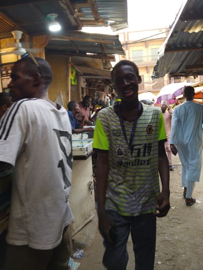 The image captures a vibrant, bustling street market scene in Tarauni, Nigeria, under natural daylight. Makeshift stalls with corrugated metal roofs and wooden supports line the area, suggesting an outdoor or semi-outdoor commercial space. A multi-story, light-colored building is visible in the background through an opening in the market structures.

In the foreground, a young man with dark skin stands centrally, facing the camera with a broad smile or grin. He is wearing a distinctive short-sleeved t-shirt with a patterned design of lime green and grey-black stripes. The shirt features a "WOLVES" logo (a stylized wolf's head), Chinese characters "万博" (Wàn Bó), and the English text "ManBet." A dark lanyard hangs around his neck. He is dressed in dark trousers.

To the left, partially obscured, an older man stands with his back mostly to the camera, wearing a light grey t-shirt with black stripes on the sleeves and a large black number "7" printed on the back. He has sunglasses perched on his head.

Several other individuals populate the mid-ground and background. On the right, a man in a light blue traditional robe (likely a babanriga or kaftan) walks away from the camera, holding a mobile phone. Behind the central subject, another man in a light pinkish-brown traditional garment, wearing glasses, can be seen. Further right, a man with a beard leans on a white display case, suggesting a vendor or shopkeeper. Adjacent to the display case is a diamond-plate textured metal surface, possibly a locked storage box or door.

Overhead, a bright white LED light bulb and a ceiling fan are attached to the market's roof structure. In the background, large purple and lighter-colored umbrellas provide additional shade. The ground is a dusty, unpaved surface with visible litter, including plastic wrappers. The scene conveys a busy, active market environment.
